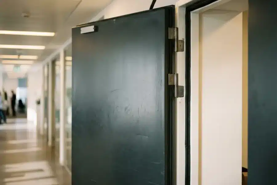 Stalen brandwerende deur met verstevigde scharnieren staat op een kier in moderne kantoorgang, met intumescerende afdichtingen zichtbaar langs de framerand
