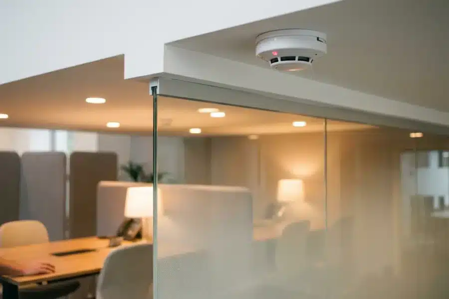 Moderne rookmelder met rode LED-indicator aan wit plafond boven glazen scheidingswand in professioneel kantoor