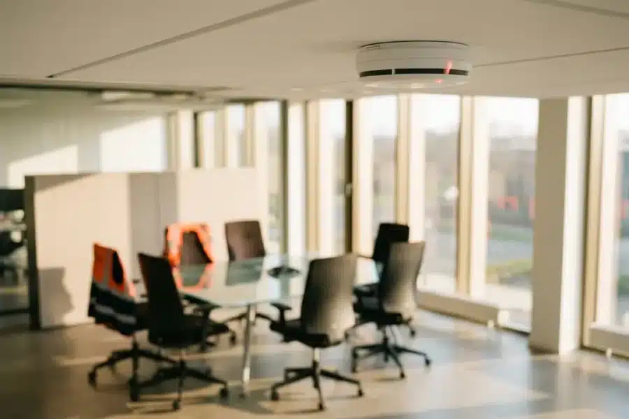 Witte rookmelder met rode LED-indicator gemonteerd aan kantoorplafond boven moderne werkruimte met glazen tafel